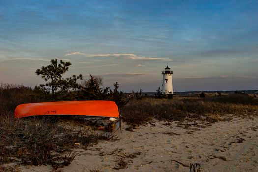 View of the Edgartown Harbor Light and an orange canoe overturned on a sandy beach with coastal vegetation under a twilight sky in Edgartown Massachusetts United States.