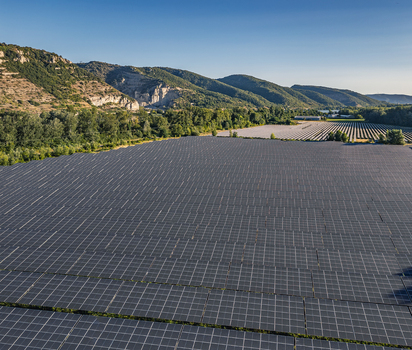 Aerial view of solar photovoltaic panels forming geometric rows at a utility scale solar farm set against rolling hills and lush green trees in Le Pouzin Auvergne Rhone Alpes France. Aerial view of solar photovoltaic panels forming geometric rows at a utility scale solar farm set against rolling hills and lush green trees in Le Pouzin Auvergne Rhone Alpes France.
