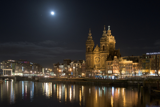 View of the illuminated facade of the Basilica of Saint Nicholas mirrored in the still waters under a bright moon Amsterdam North Holland Netherlands.