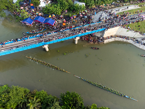 Aerial view of vibrant boat races cutting through the murky river under a crowded bridge where spectators gather Gopalganj Dhaka Division Bangladesh. Aerial view of vibrant boat races cutting through the murky river under a crowded bridge where spectators gather Gopalganj Dhaka Division Bangladesh.