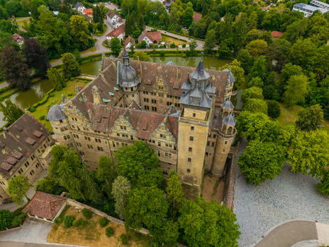 Aerial view of the historic Neuenstein Castle with stone walls tiled roofs and moat set amidst lush green trees and parkland in Neuenstein Baden Wurttemberg Germany. Aerial view of the historic Neuenstein Castle with stone walls tiled roofs and moat set amidst lush green trees and parkland in Neuenstein Baden Wurttemberg Germany.