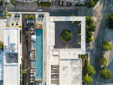 Aerial view of a modern hotel building featuring a long rectangular swimming pool sun loungers and a circular courtyard with a central tree in Merida Yucatan Mexico. Aerial view of a modern hotel building featuring a long rectangular swimming pool sun loungers and a circular courtyard with a central tree in Merida Yucatan Mexico.