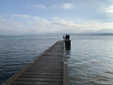 Japan 06 July 2026 View of three people standing at the end of a wooden pier extending into a calm lake with distant mountains under a cloudy sky. Japan 06 July 2026 View of three people standing at the end of a wooden pier extending into a calm lake with distant mountains under a cloudy sky.