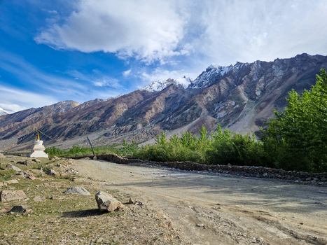 View of the Penzi La road winding through the rugged landscape with a traditional stupa snow capped mountains and lush green trees under a bright blue sky in Zanskar Ladakh India.