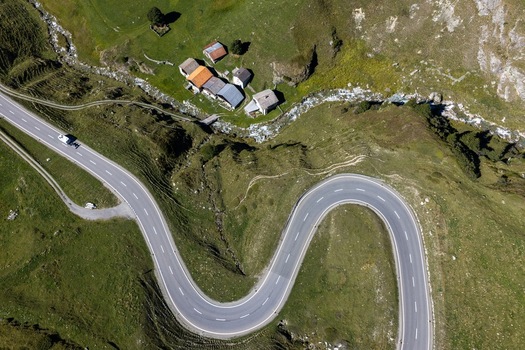 Aerial view of the winding Julier Pass road surrounded by lush green alpine meadows a small cluster of mountain buildings and a flowing stream in Surses Grisons Switzerland. Aerial view of the winding Julier Pass road surrounded by lush green alpine meadows a small cluster of mountain buildings and a flowing stream in Surses Grisons Switzerland.