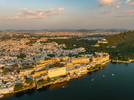 Aerial view of the City Palace complex on the banks of Lake Pichola with historic architecture rooftops and lush hills under golden hour light in Udaipur Rajasthan India.