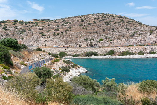 View of the turquoise sea bay surrounded by rocky arid hills with sparse green shrubs and a coastal road in Saronida Attica Greece.