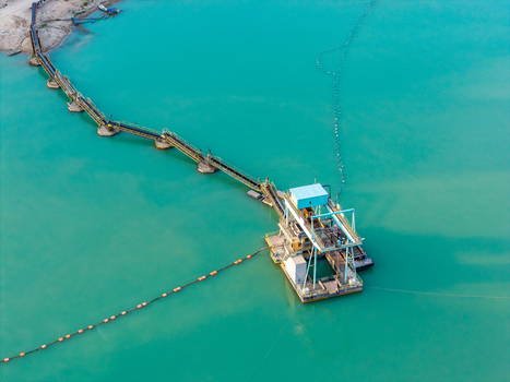 Aerial view of a large scale sand and gravel mine with a floating processing platform and a long conveyor belt system extending across turquoise water in Poland.