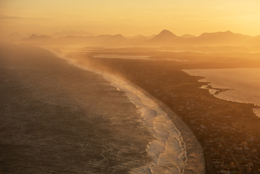 Aerial view of the coastline at sunset with golden light reflecting on the ocean waves and distant mountains in Rio de Janeiro Rio de Janeiro Brazil. Aerial view of the coastline at sunset with golden light reflecting on the ocean waves and distant mountains in Rio de Janeiro Rio de Janeiro Brazil.