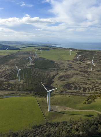 Aerial view of a windfarm with white turbines scattered across rolling hills and green fields near the coast under a blue sky with clouds Scotland United Kingdom. Aerial view of a windfarm with white turbines scattered across rolling hills and green fields near the coast under a blue sky with clouds Scotland United Kingdom.