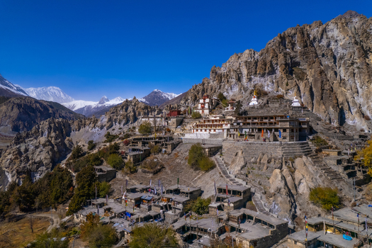 Aerial view of the historic Jharkot Monastery and village nestled against rugged rocky Himalayan cliffs under a clear blue sky in Nepal.