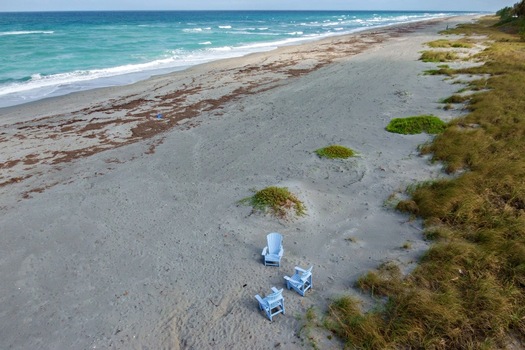 Aerial view of azure waters meeting the sandy shore dotted with beach vegetation and three inviting chairs Jupiter Island Florida United States. Aerial view of azure waters meeting the sandy shore dotted with beach vegetation and three inviting chairs Jupiter Island Florida United States.