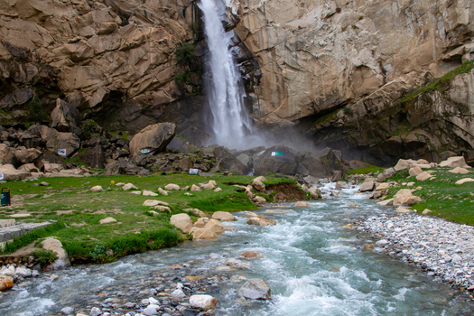 View of Khamosh Waterfall cascading down steep rocky cliffs into a clear mountain stream surrounded by green grass and boulders in Skardu Gilgit Pakistan.