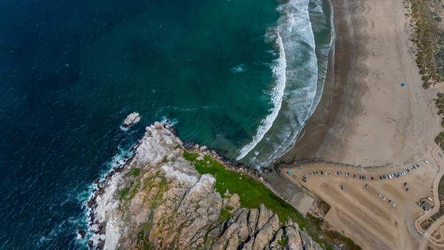 Aerial view of Morro Rock featuring rugged cliffs turquoise ocean waves crashing against the shore and a sandy beach with a parking area Morro Bay California United States.