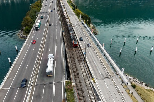 Aerial view of a train traveling on tracks alongside a busy highway with cars and trucks on a causeway over Lake Lugano in Melide Ticino Switzerland. Aerial view of a train traveling on tracks alongside a busy highway with cars and trucks on a causeway over Lake Lugano in Melide Ticino Switzerland.