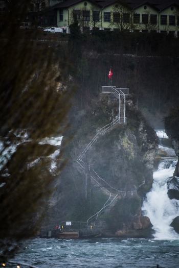 View of a powerful waterfall cascading down the rocks mist swirling around the Swiss flag atop a stone perch Neuhausen am Rheinfall Switzerland. View of a powerful waterfall cascading down the rocks mist swirling around the Swiss flag atop a stone perch Neuhausen am Rheinfall Switzerland.
