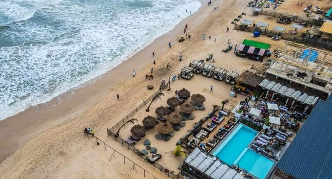 Aerial view of a beach resort featuring a swimming pool thatched umbrellas and lounge areas with people relaxing on the sandy shore in Ghana. Aerial view of a beach resort featuring a swimming pool thatched umbrellas and lounge areas with people relaxing on the sandy shore in Ghana.