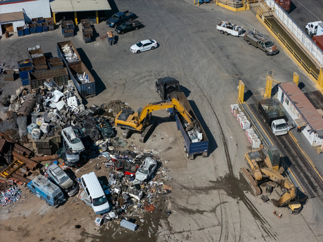 Aerial view of an industrial scrap yard in Carson California United States featuring yellow excavators loading scrap metal into trucks amidst piles of discarded vehicles.