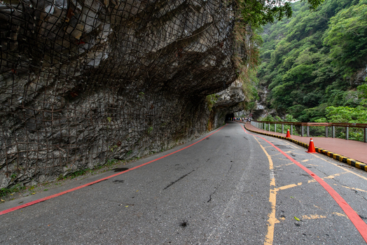 View of Taroko National Park featuring a road winding through a rock tunnel with safety netting red traffic cones and a wooden pedestrian walkway in Xiulin Township Hualien County Taiwan.