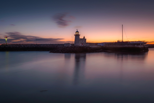 View of Howth Harbour lighthouse at sunset with calm water reflections and a stone pier under a colorful sky in Ireland. View of Howth Harbour lighthouse at sunset with calm water reflections and a stone pier under a colorful sky in Ireland.