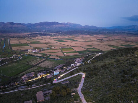 Aerial view of the agricultural landscape with a winding road illuminated by car lights at dusk set against a backdrop of mountains in Saranda Vlore County Albania. Aerial view of the agricultural landscape with a winding road illuminated by car lights at dusk set against a backdrop of mountains in Saranda Vlore County Albania.
