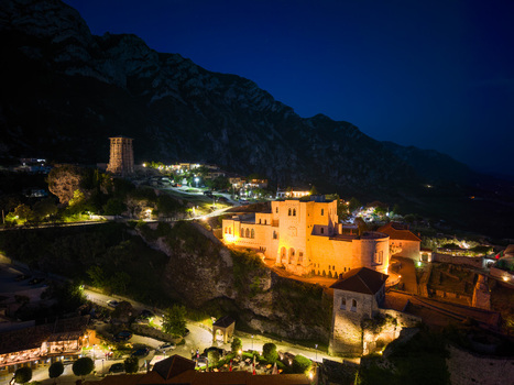 Aerial view of the illuminated Kruje Castle and its historic clock tower perched on rocky cliffs against dark mountains at night in Kruje Durres County Albania.