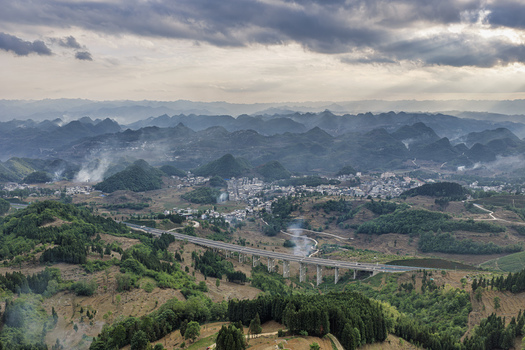 Aerial view of a long highway bridge spanning a valley near a rural village and karst mountains under a dramatic cloudy sky in Gaoxing Village Guizhou China.