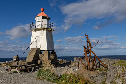 View of Laukvik fyr lighthouse on a rocky shore with a rusty metal sculpture and a picnic table under a blue sky Laukvik Nordland Norway. View of Laukvik fyr lighthouse on a rocky shore with a rusty metal sculpture and a picnic table under a blue sky Laukvik Nordland Norway.
