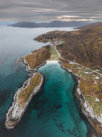 Aerial view of the rugged coastline of Senja with turquoise waters rocky cliffs a small beach and scattered houses along the road in Senja Troms Norway.