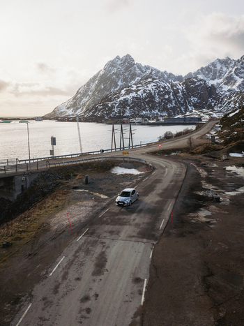 Aerial view of a lone vehicle traversing a road leading to the Kåkernbrua bridge against a backdrop of snow dusted mountains Kåkernbrua Nordland Norway. Aerial view of a lone vehicle traversing a road leading to the Kåkernbrua bridge against a backdrop of snow dusted mountains Kåkernbrua Nordland Norway.