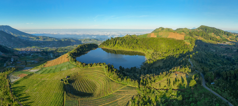 Aerial view of Telaga Dringo lake surrounded by lush green hills terraced agricultural fields and a distant village under a clear blue sky Kecamatan Batur Jawa Tengah Indonesia.