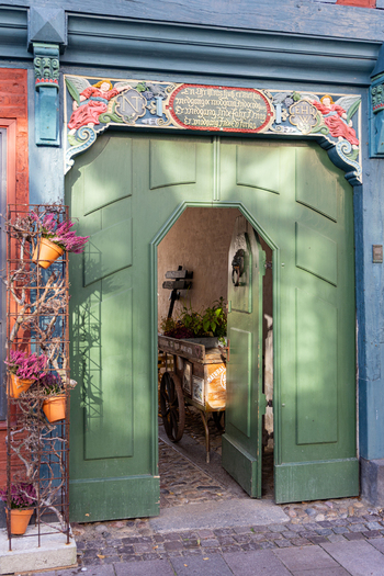 View of an ornate historic green wooden door with carved angels and inscriptions leading to a courtyard with a wooden cart Ribe Denmark. View of an ornate historic green wooden door with carved angels and inscriptions leading to a courtyard with a wooden cart Ribe Denmark.
