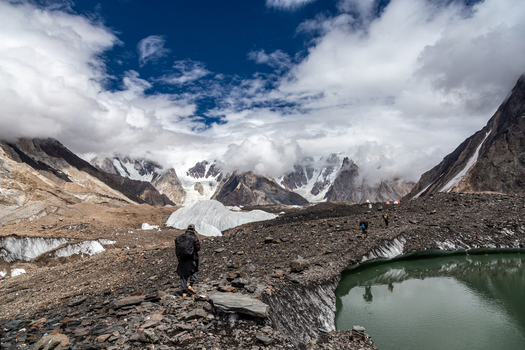 View of a hiker walking along the rocky moraine and glacial lake of the Baltoro Glacier surrounded by snow capped peaks under a cloudy sky in Baltoro Gilgit Baltistan Pakistan.