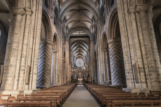 View of the interior of Durham Cathedral featuring massive Norman columns with chevron carvings a long nave with wooden pews and a rose window in Durham England United Kingdom. View of the interior of Durham Cathedral featuring massive Norman columns with chevron carvings a long nave with wooden pews and a rose window in Durham England United Kingdom.