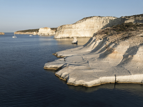 Aerial view of the white volcanic rock formations of Sarakiniko beach with calm blue sea and sailboats in the distance in Milos Greece. Aerial view of the white volcanic rock formations of Sarakiniko beach with calm blue sea and sailboats in the distance in Milos Greece.