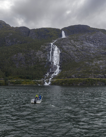 Aerial view of Langfossen waterfall cascading down steep rocky cliffs into the fjord with a small boat and people on the water in Fjaera Vestland Norway. Aerial view of Langfossen waterfall cascading down steep rocky cliffs into the fjord with a small boat and people on the water in Fjaera Vestland Norway.