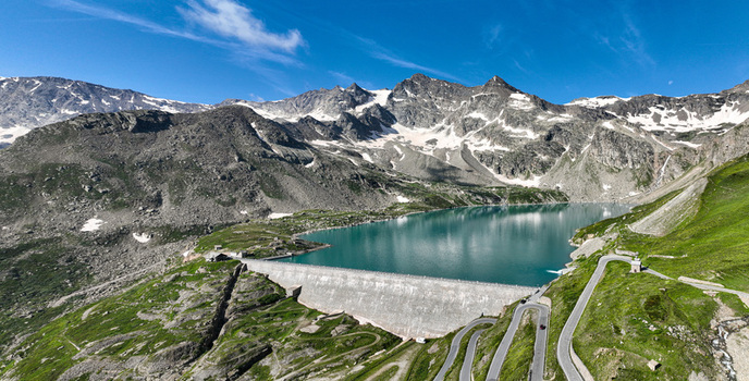 Aerial view of Serru Lake and its massive dam surrounded by rugged alpine peaks with snow patches and winding mountain roads in Ceresole Reale Piedmont Italy. Aerial view of Serru Lake and its massive dam surrounded by rugged alpine peaks with snow patches and winding mountain roads in Ceresole Reale Piedmont Italy.