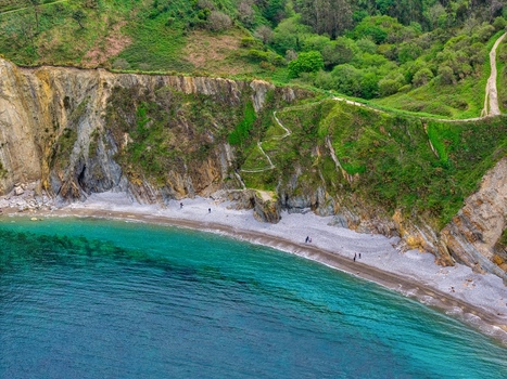 Aerial view of Playa del Silencio with turquoise water steep rocky cliffs lush green hills and people walking on the pebble shore Cudillero Asturias Spain.