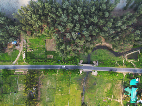 Aerial view of a straight asphalt road with a white car crossing a small stream between dense green trees and agricultural fields in Cox s Bazar Chittagong Division Bangladesh. Aerial view of a straight asphalt road with a white car crossing a small stream between dense green trees and agricultural fields in Cox s Bazar Chittagong Division Bangladesh.