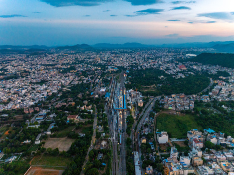 Aerial view of Udaipur Railway Station with its platforms and tracks cutting through the dense urban landscape surrounded by lush greenery and distant hills in Udaipur Rajasthan India.