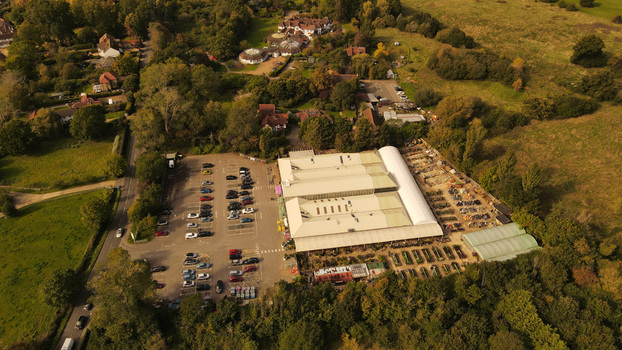 Aerial view of a garden center with a large greenhouse a parking lot filled with cars and outdoor plant displays surrounded by trees and green fields England United Kingdom. Aerial view of a garden center with a large greenhouse a parking lot filled with cars and outdoor plant displays surrounded by trees and green fields England United Kingdom.