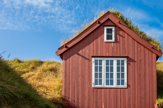 View of a traditional red wooden house with a grass roof set against a clear blue sky in the Faroe Islands. View of a traditional red wooden house with a grass roof set against a clear blue sky in the Faroe Islands.