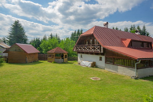 View of a red roofed chalet basks in the sunlit yard lush green grass contrasting against the backdrop of a forest under a bright sky Donovaly Slovakia. View of a red roofed chalet basks in the sunlit yard lush green grass contrasting against the backdrop of a forest under a bright sky Donovaly Slovakia.