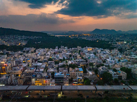 Aerial view of the cityscape of Udaipur at sunset with dense urban buildings illuminated streets and distant hills under a dramatic cloudy sky in Udaipur Rajasthan India. Aerial view of the cityscape of Udaipur at sunset with dense urban buildings illuminated streets and distant hills under a dramatic cloudy sky in Udaipur Rajasthan India.
