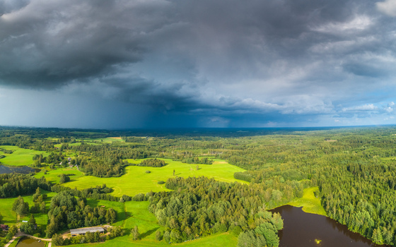 Aerial view of a rural landscape with lush green fields dense forests and a small lake under dramatic dark storm clouds in Latvia. Aerial view of a rural landscape with lush green fields dense forests and a small lake under dramatic dark storm clouds in Latvia.