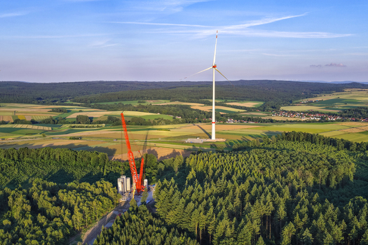 Aerial view of a wind turbine and a large red construction crane situated in a dense forest near agricultural fields in Hofgeismar Hessen Germany.