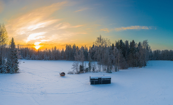 Aerial view of a snowy landscape featuring a small cabin and a barrel sauna nestled near a forest during a vibrant sunset in Latvia. Aerial view of a snowy landscape featuring a small cabin and a barrel sauna nestled near a forest during a vibrant sunset in Latvia.