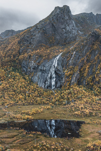 Aerial view of a waterfall cascading down a rocky mountain slope in Lofoten surrounded by vibrant autumn foliage and a reflective pond in Leknes Nordland Norway. Aerial view of a waterfall cascading down a rocky mountain slope in Lofoten surrounded by vibrant autumn foliage and a reflective pond in Leknes Nordland Norway.