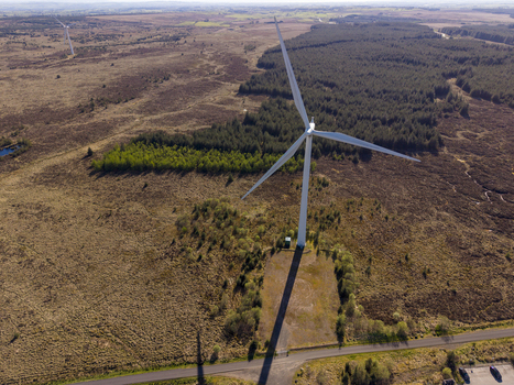 Aerial view of a large white wind turbine standing in a vast rural landscape with dense forests and open fields under a clear sky.