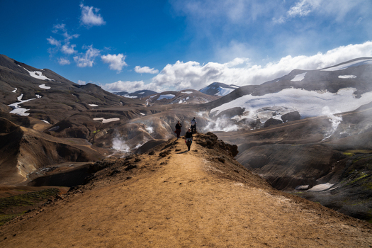 View of a rugged otherworldly landscape with billowing white steam rising from the earth as hikers trek across the brown terrain Hrunamannahreppur Iceland.